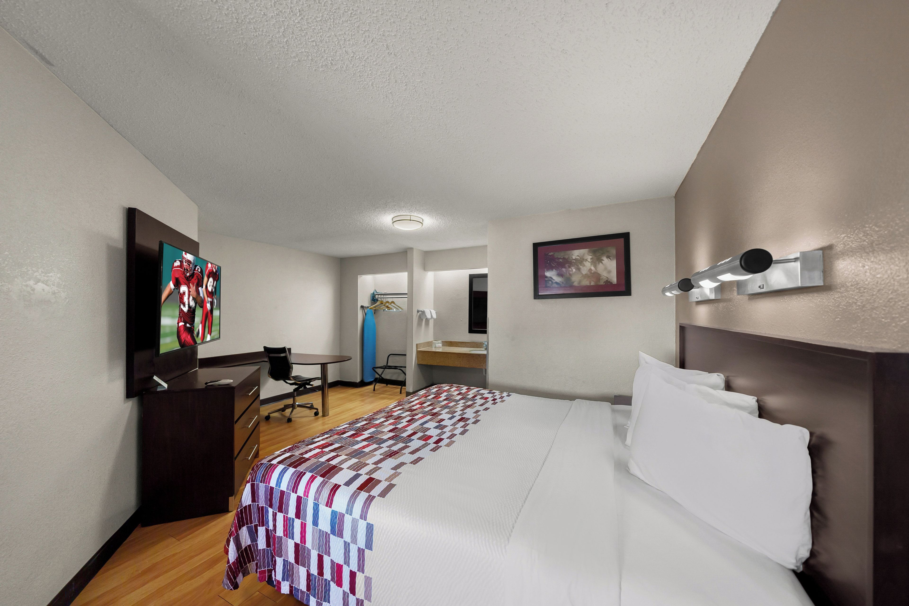 Habitación de hotel con cama, escritorio, silla, televisor, un cuadro y baño visible a través de una puerta abierta.