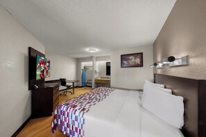 Habitación de hotel con cama, escritorio, silla, televisor, un cuadro y baño visible a través de una puerta abierta.