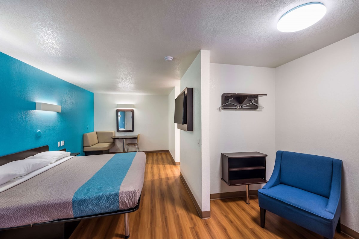 Una habitación de hotel con una cama, una silla azul, un televisor, una mesita y un espejo en la pared.