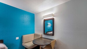 A small room with a blue wall, a beige sofa, a round table with a chair, and a wall-mounted mirror. - Motel 6 Irving, TX - Irving DFW Airport East (Irving)