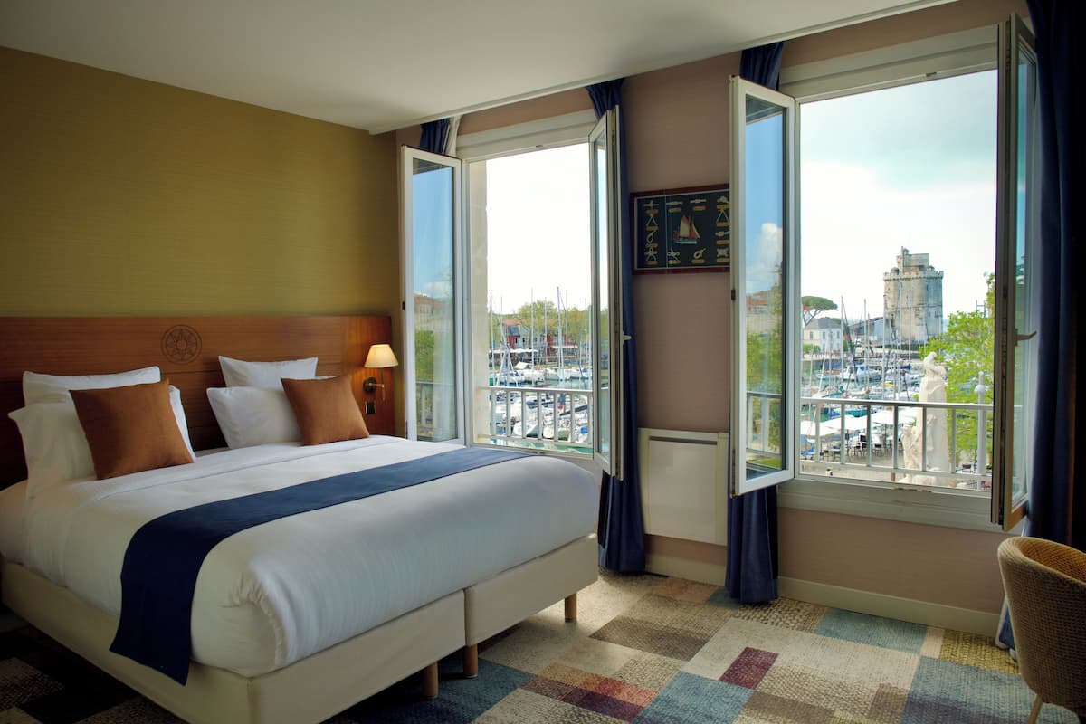 A hotel room with a large bed, a view of a marina, and a cityscape visible through the windows.