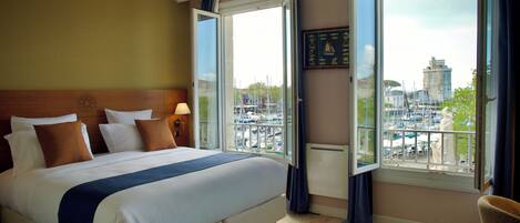 A hotel room with a large bed, a view of a marina, and a cityscape visible through the windows.