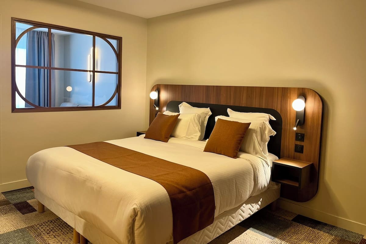 A hotel room with a large bed, a bedside table, a nightstand, a window with curtains, and a wall-mounted light fixture.