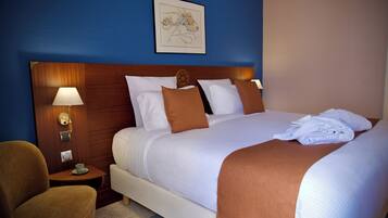 Habitación de hotel con una cama grande, una mesita de noche con lámpara, una silla e imagen enmarcadas en la pared.