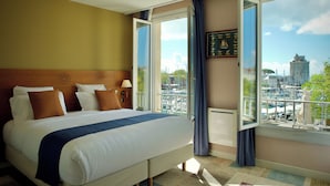 Un cuarto de hotel con cama, ventana con cortinas, vista a la ciudad y un cuadro en la pared.