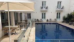 Piscina con barrera de vidrio, sillas de descanso y mesas con sombrillas.