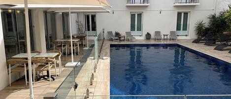 A swimming pool with a glass barrier, lounge chairs, and tables with umbrellas.
