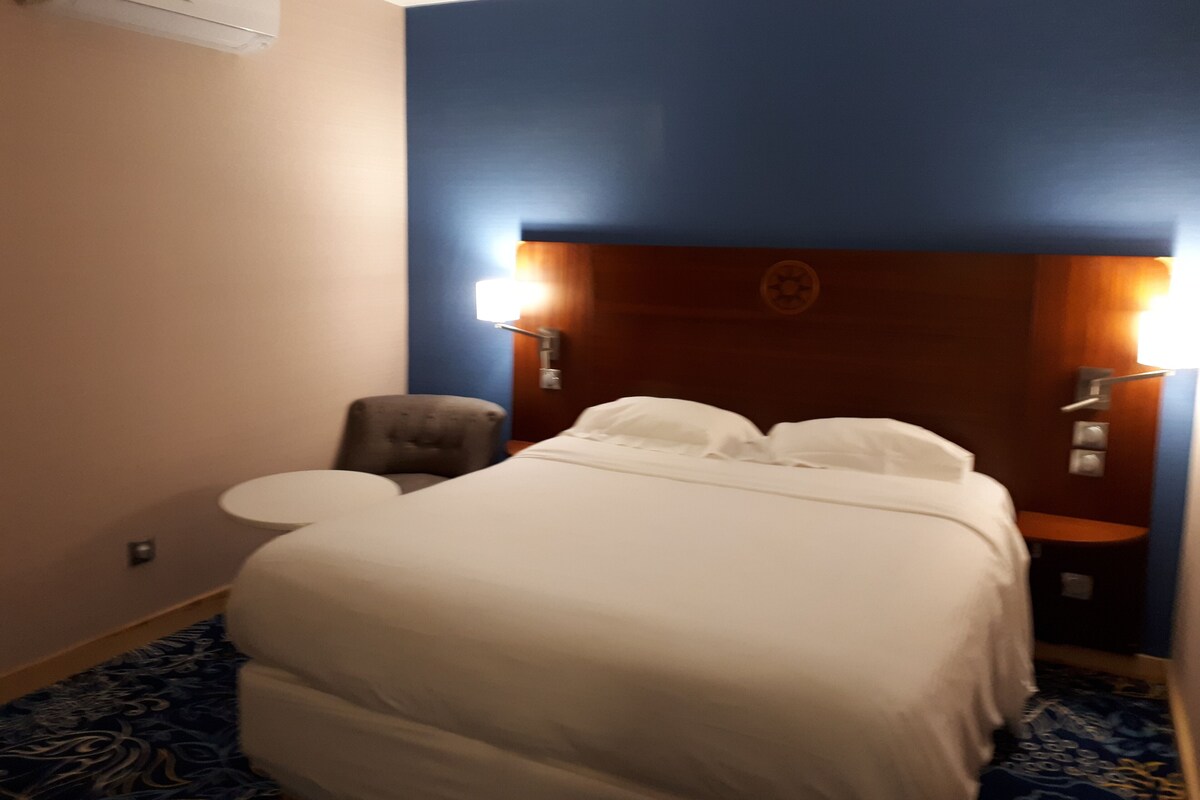 A hotel room with a bed, a chair, a desk, and a wall-mounted lamp.