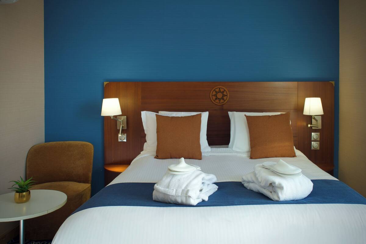 A hotel room with a bed, two pillows, a headboard, two bedside lamps, a chair, a small table with a plant, and two folded towels on the bed.