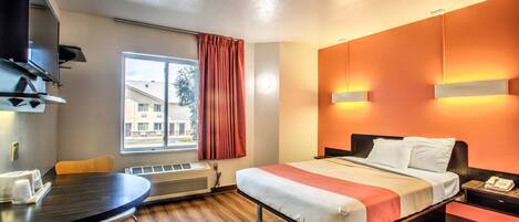 A hotel room with a bed, a desk, a chair, a TV, a window with curtains, and a view of buildings outside.