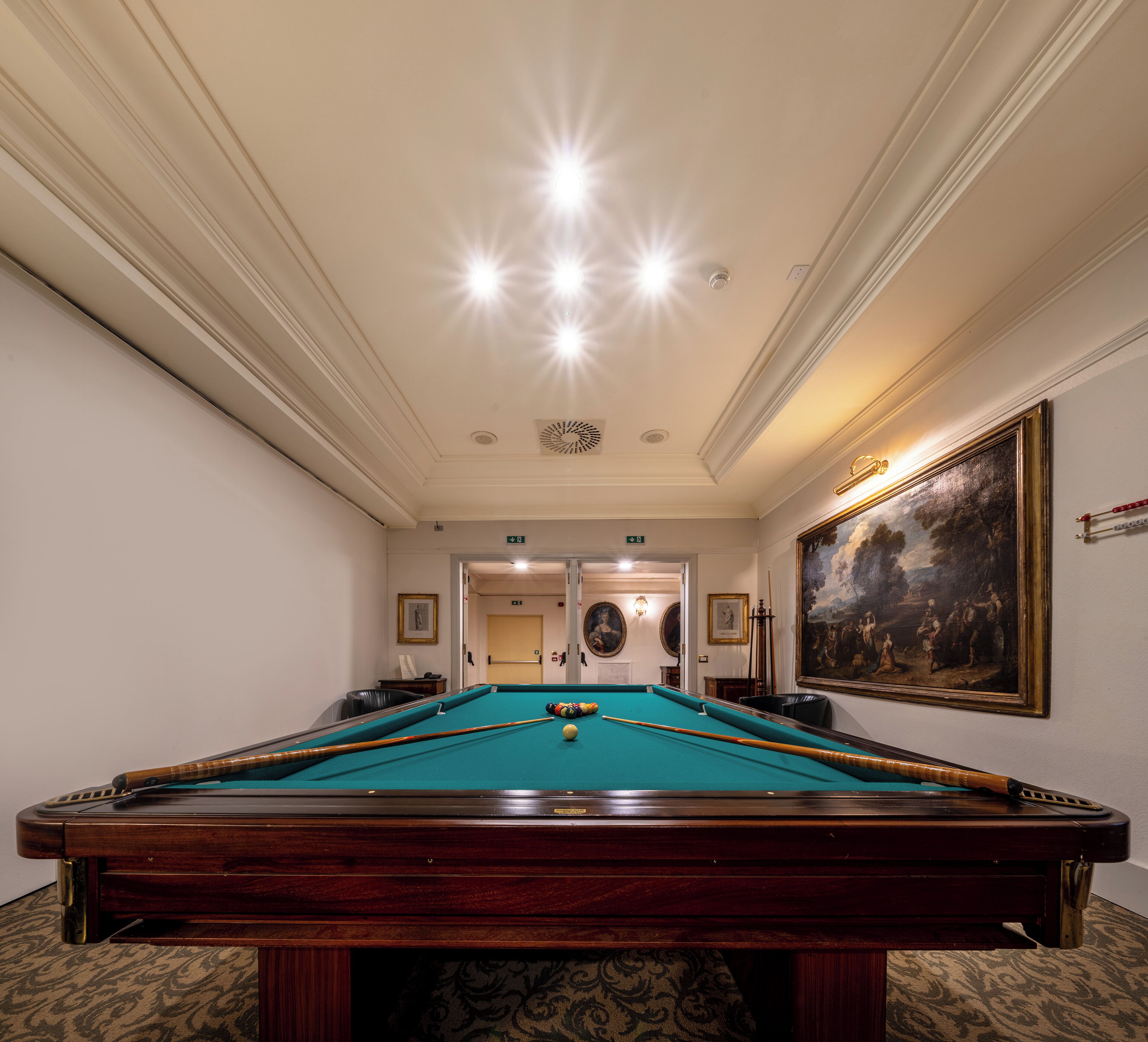 Une table de billard recouverte d’un drap vert, entourée d’un tapis à motifs. On aperçoit deux portes au fond, l’une donnant sur un couloir et l’autre sur une autre pièce.