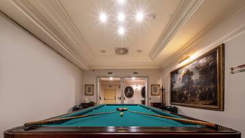 A pool table with a green felt surface, surrounded by a patterned carpet. There are two doors in the background, one leading to a hallway and the other to another room.