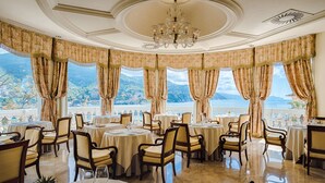 Un comedor con mesas y sillas, ventanas grandes con cortinas y un candelabro.