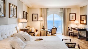 Habitación de hotel con una cama grande, un escritorio, una silla y vista al mar a través de un ventanal amplio.