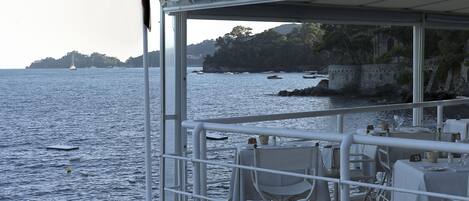 Un restaurant situé sur la côte, avec une terrasse offrant une vue sur la mer, agrémentée de tables et de chaises placées sous une auvent blanche.