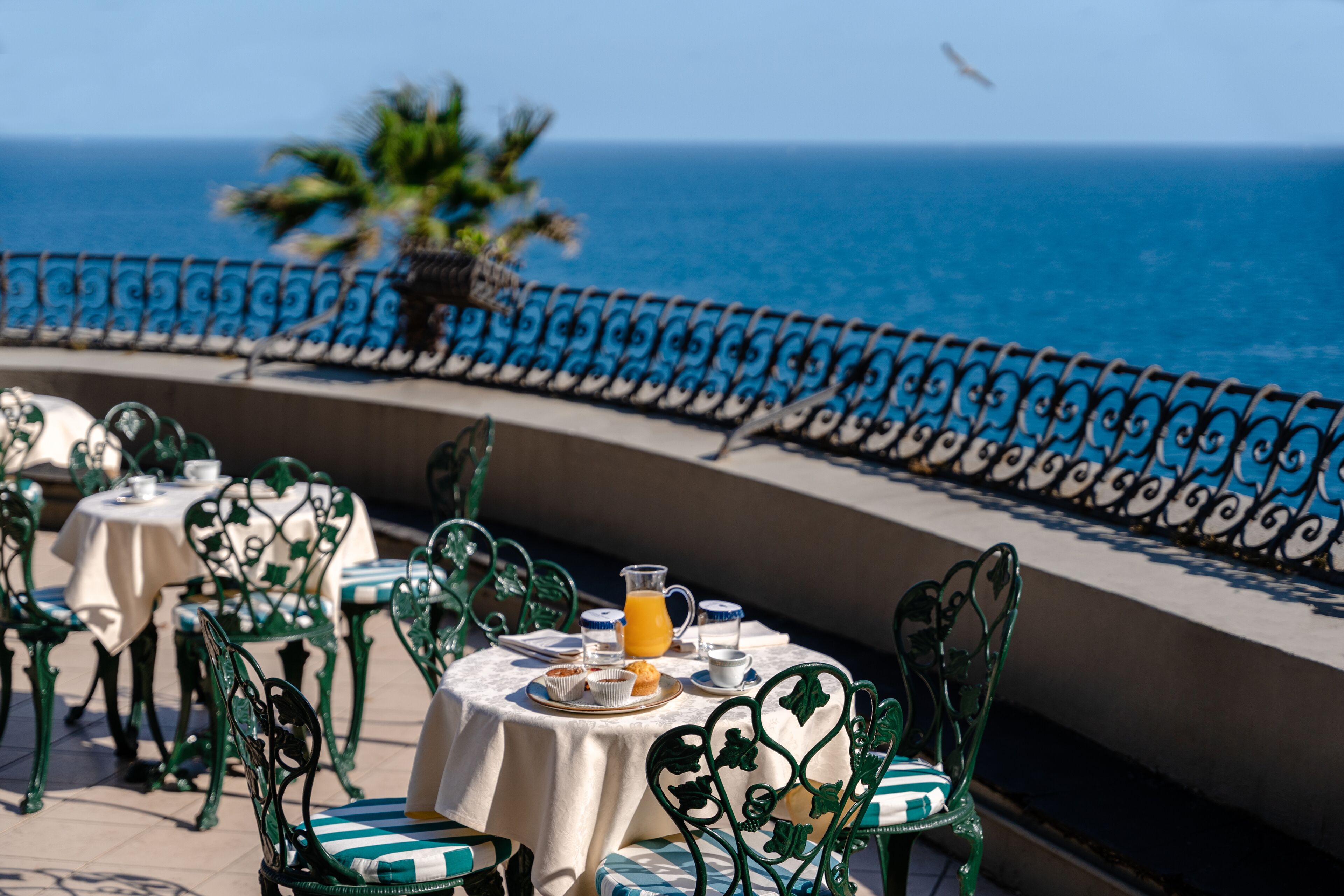 A terrace with tables and chairs, a view of the ocean, and a railing.