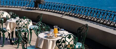 A terrace with tables and chairs, a view of the ocean, and a railing.