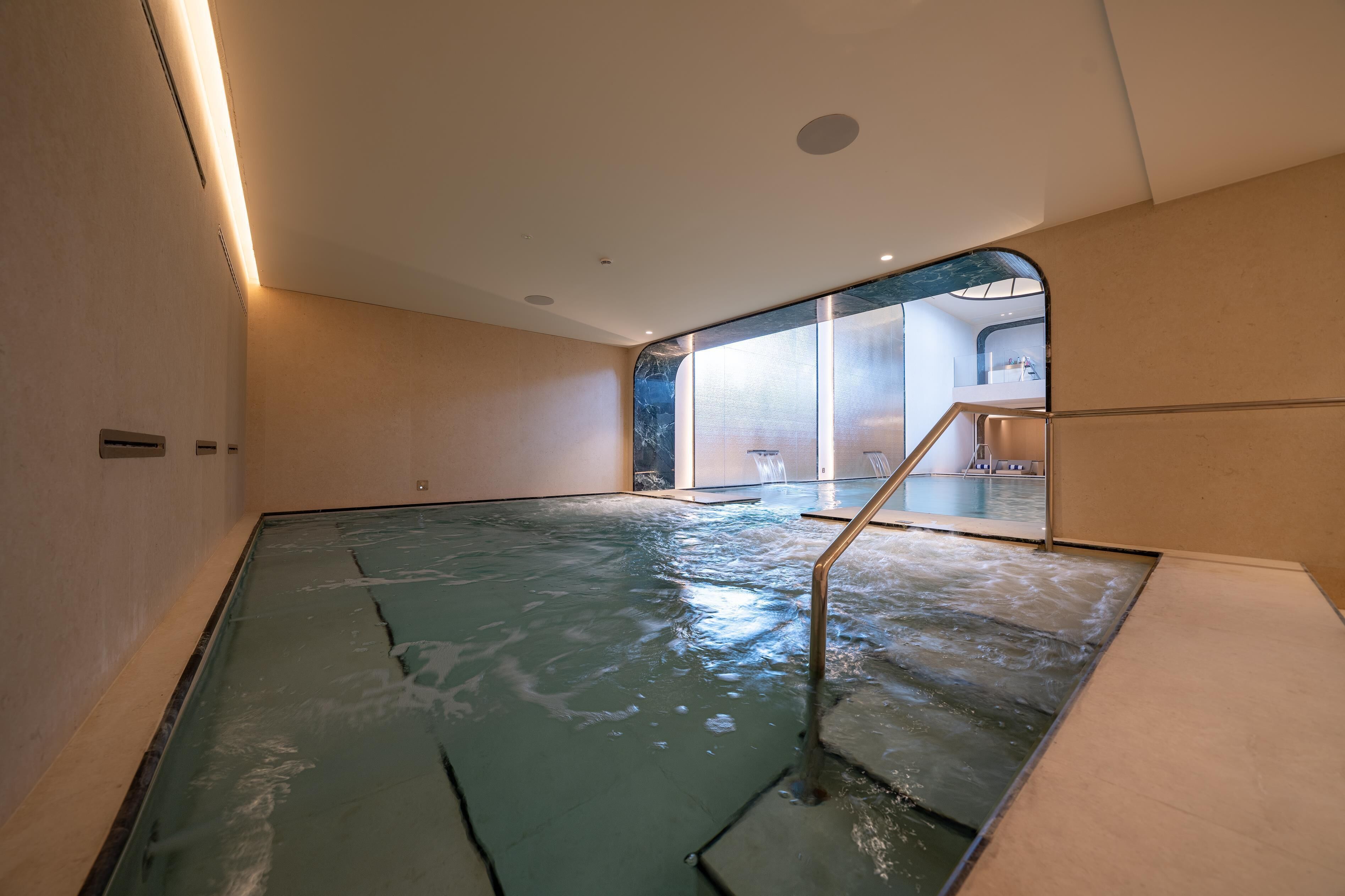 Une piscine dotée d’une paroi en verre et d’un escalier en métal, entourée de murs de couleur beige et surmontée d’un plafond avec des spots encastrés.