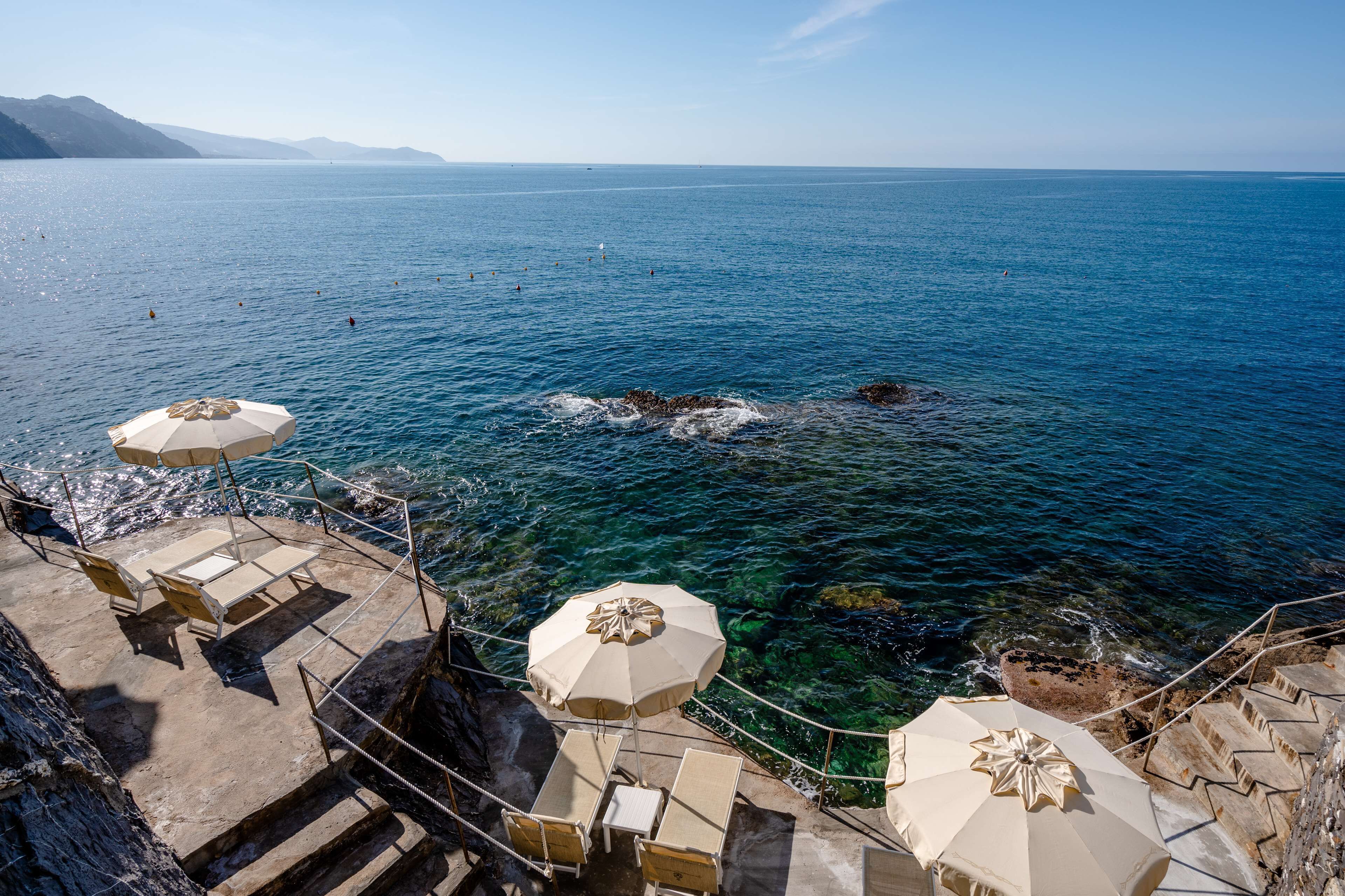 Une terrasse en bord de mer, avec une eau d’un bleu limpide, des marches en pierre et des parasols de couleur beige.