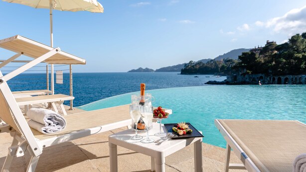 Escena junto a la piscina con sillones de descanso, una mesa con una botella, vasos y una bandeja de frutas.