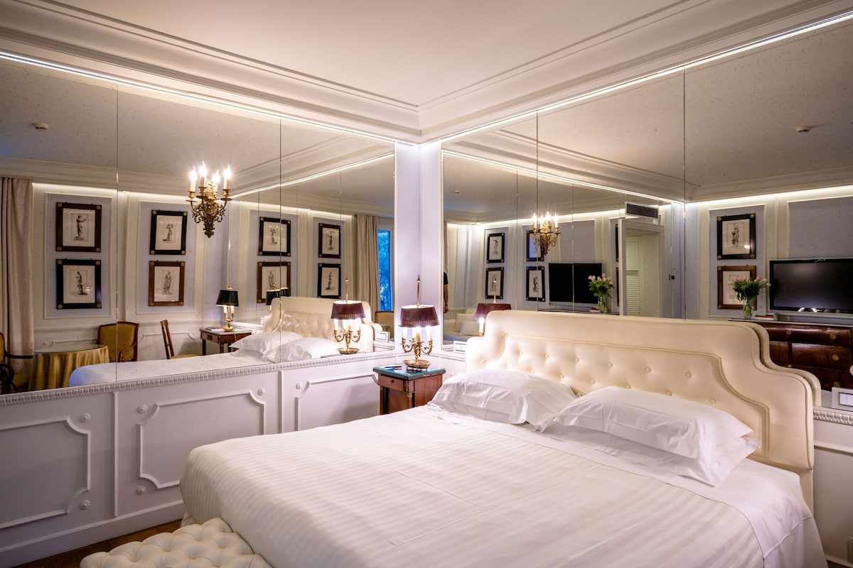 Un dormitorio lujoso con una cama grande, paredes espejadas, obras de arte enmarcadas y un candelabro.
