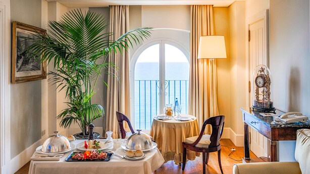 A dining area with a table set for two, a potted plant, and a view of the sea through a window. - Excelsior Palace Portofino Coast (Rapallo)