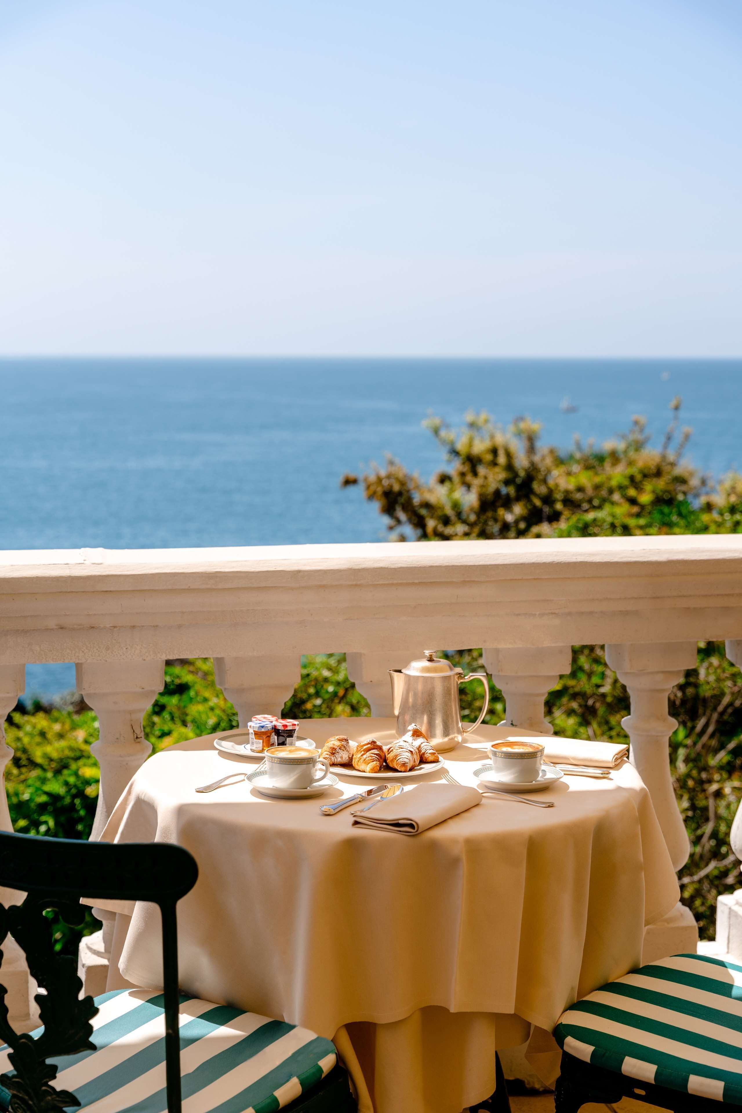 A table set for breakfast with a view of the sea, a white tablecloth, and striped cushions.
