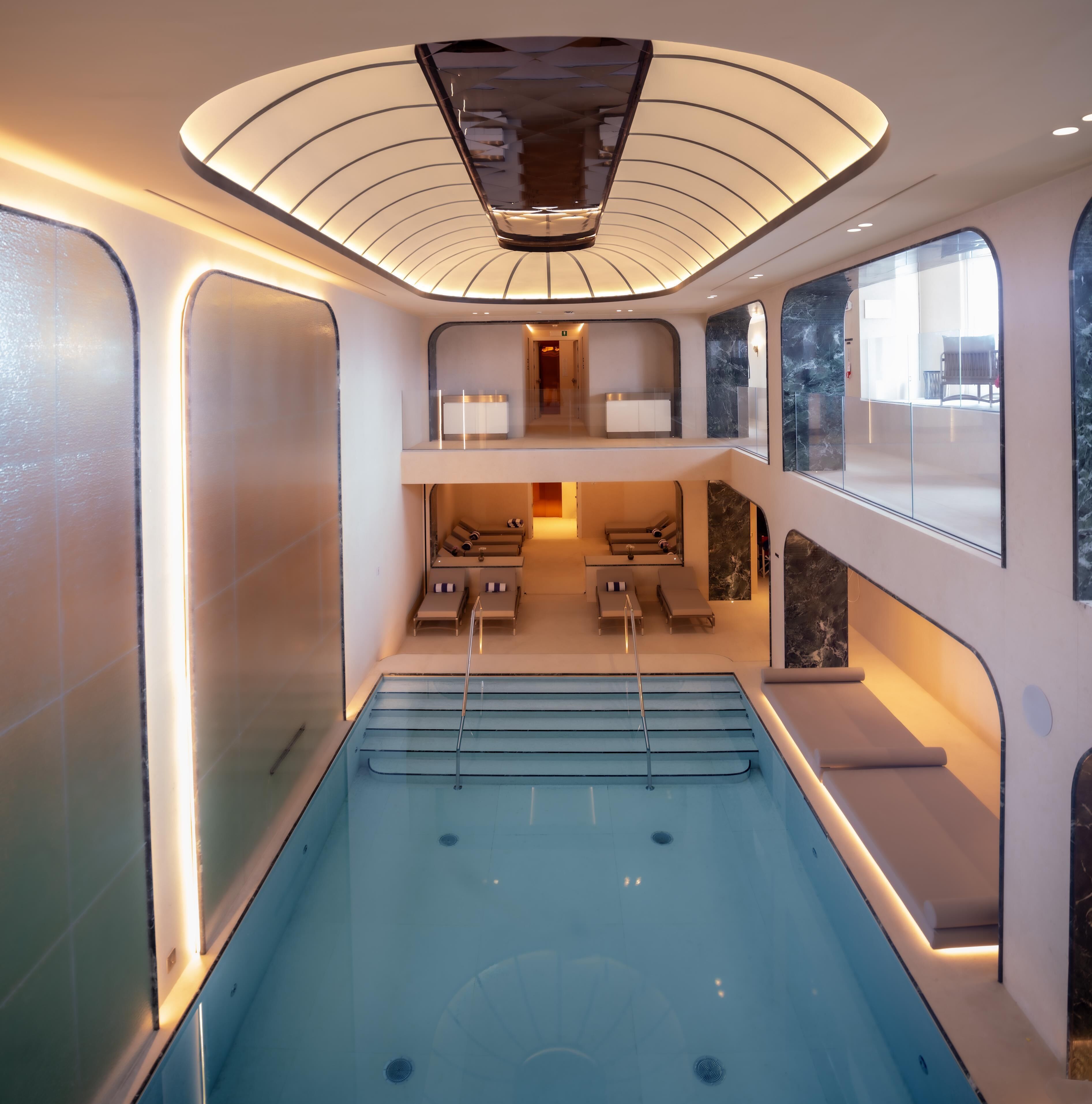A modern indoor swimming pool with a glass wall, a lounge area with seating, and a ceiling with a unique light fixture.