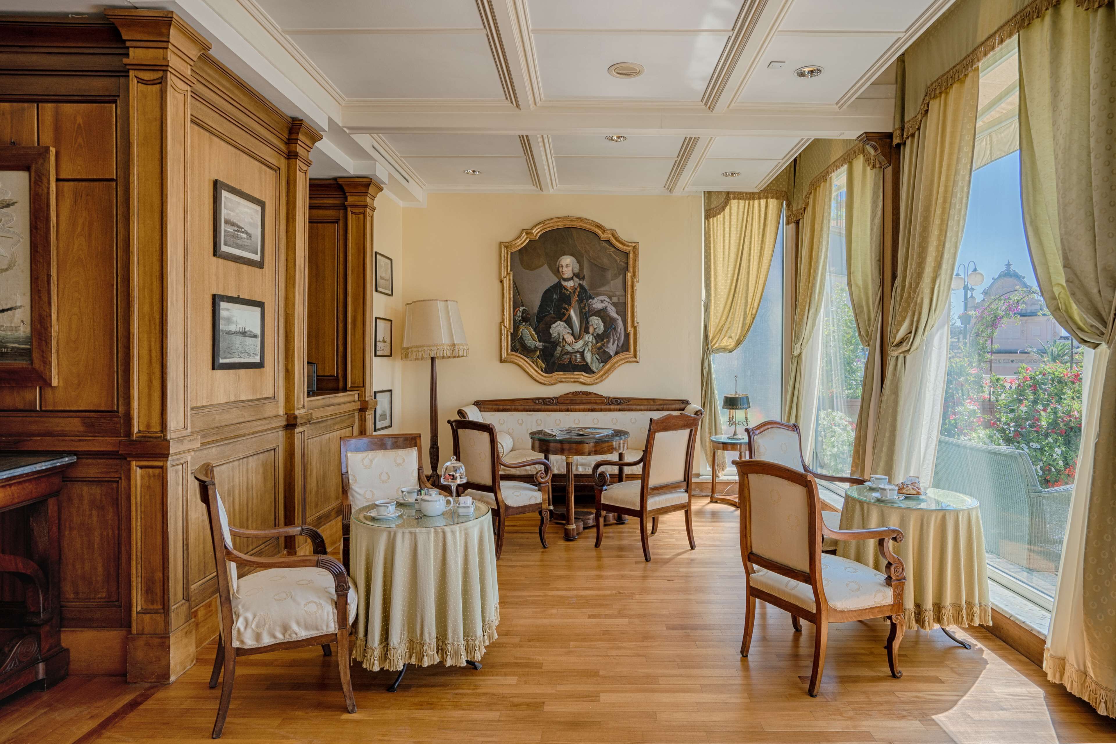 Una stanza con pannelli in legno, un grande ritratto appeso al muro, un tavolo rotondo con sedie e ampie finestre con tende.