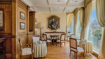 Una stanza con pannelli in legno, un grande ritratto appeso al muro, un tavolo rotondo con sedie e ampie finestre con tende.