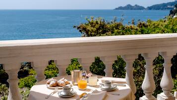 Mesa posta com croissants, sumo e café numa varanda com vista para o mar.