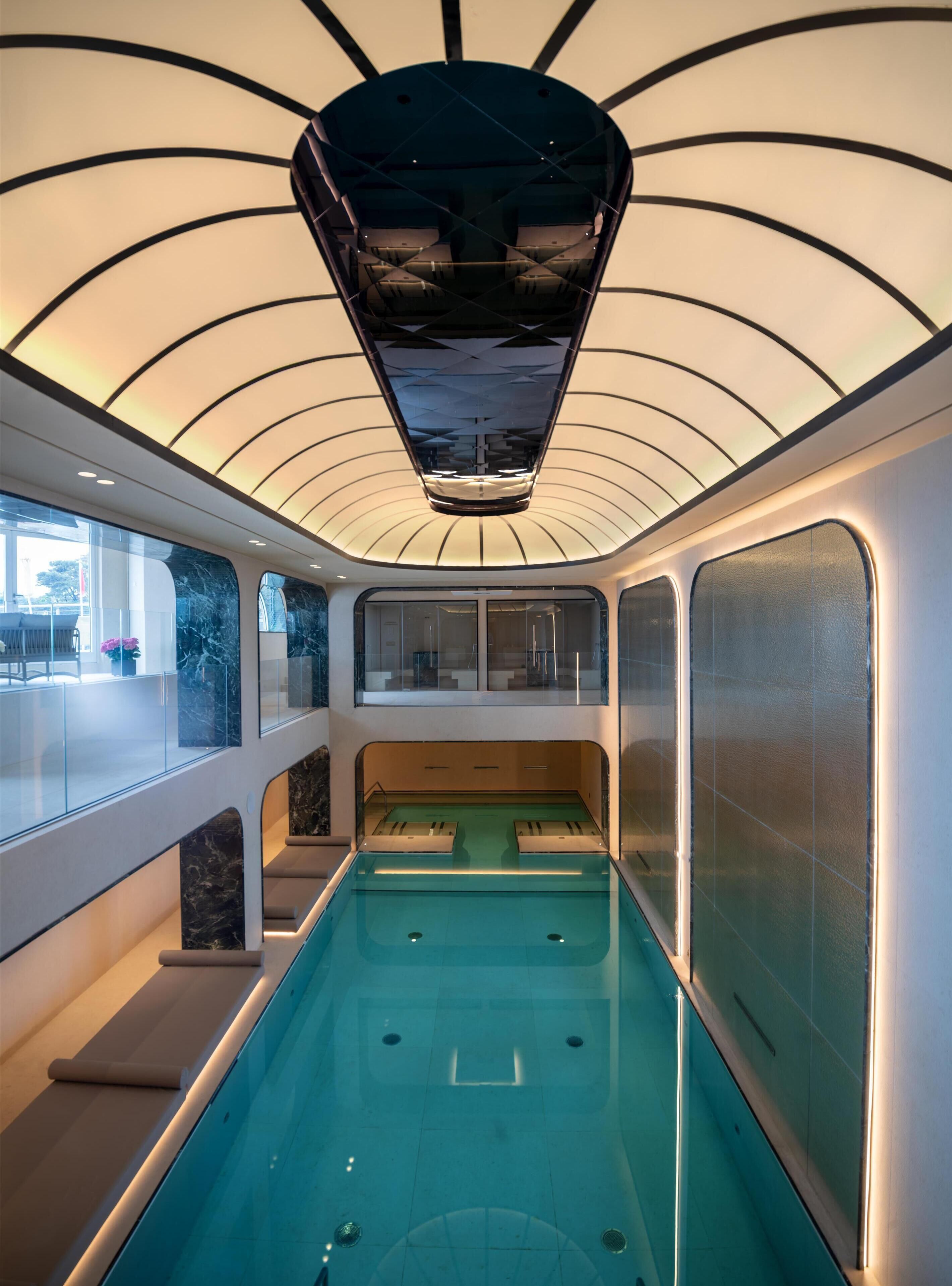 Une piscine intérieure moderne dotée d’un plafond au design unique, de murs en verre et d’un espace détente.