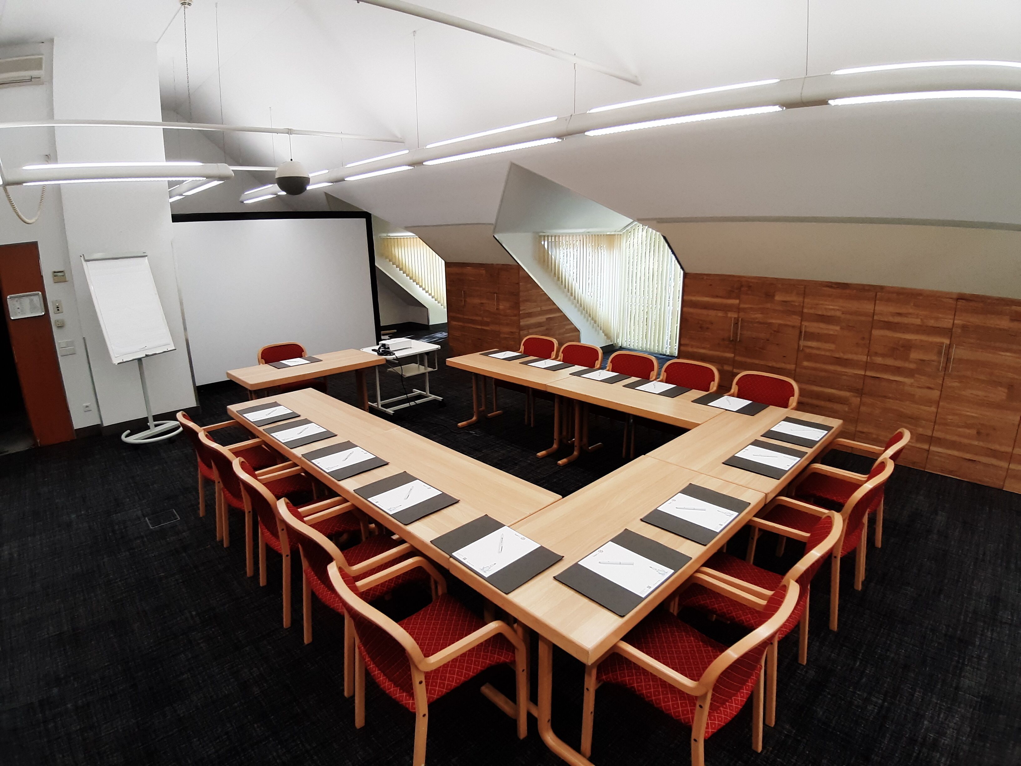Un salón de conferencias con una larga mesa, sillas rojas, una pizarra y una escalera.