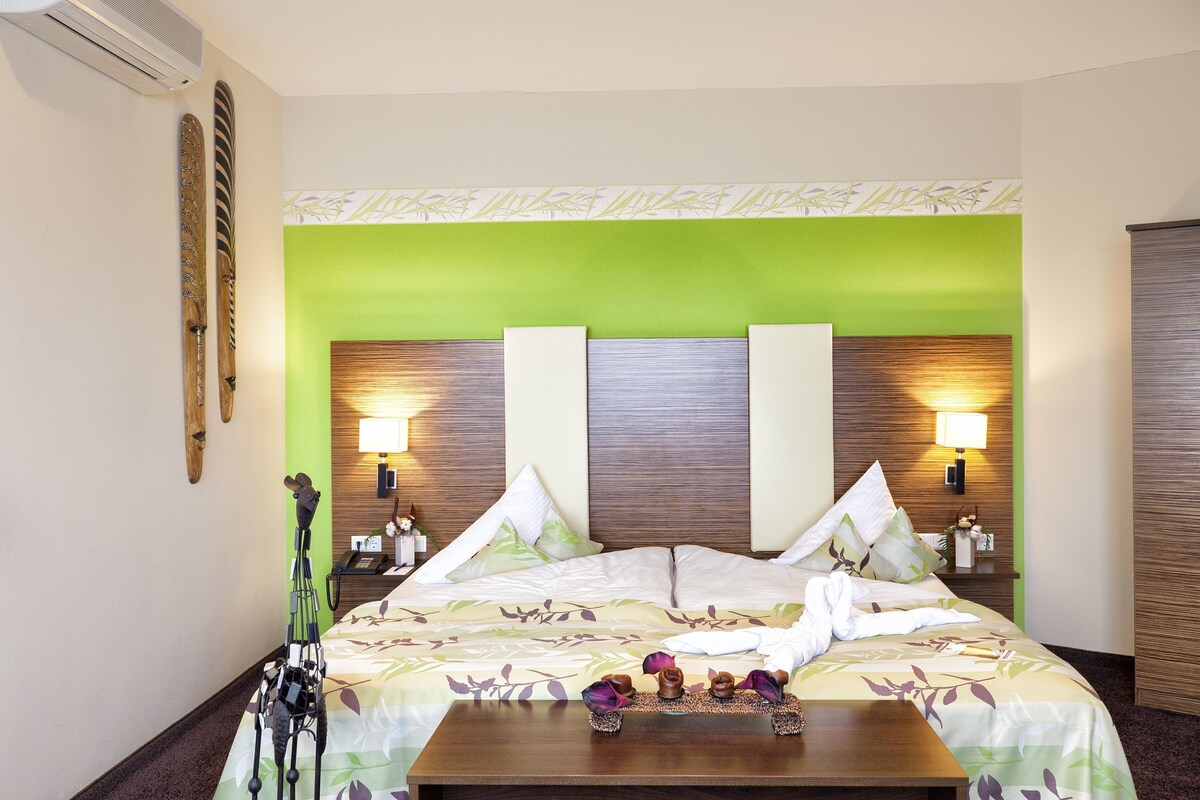 A hotel room with a bed, two bedside tables, a nightstand, a wall-mounted air conditioner, and a green accent wall.