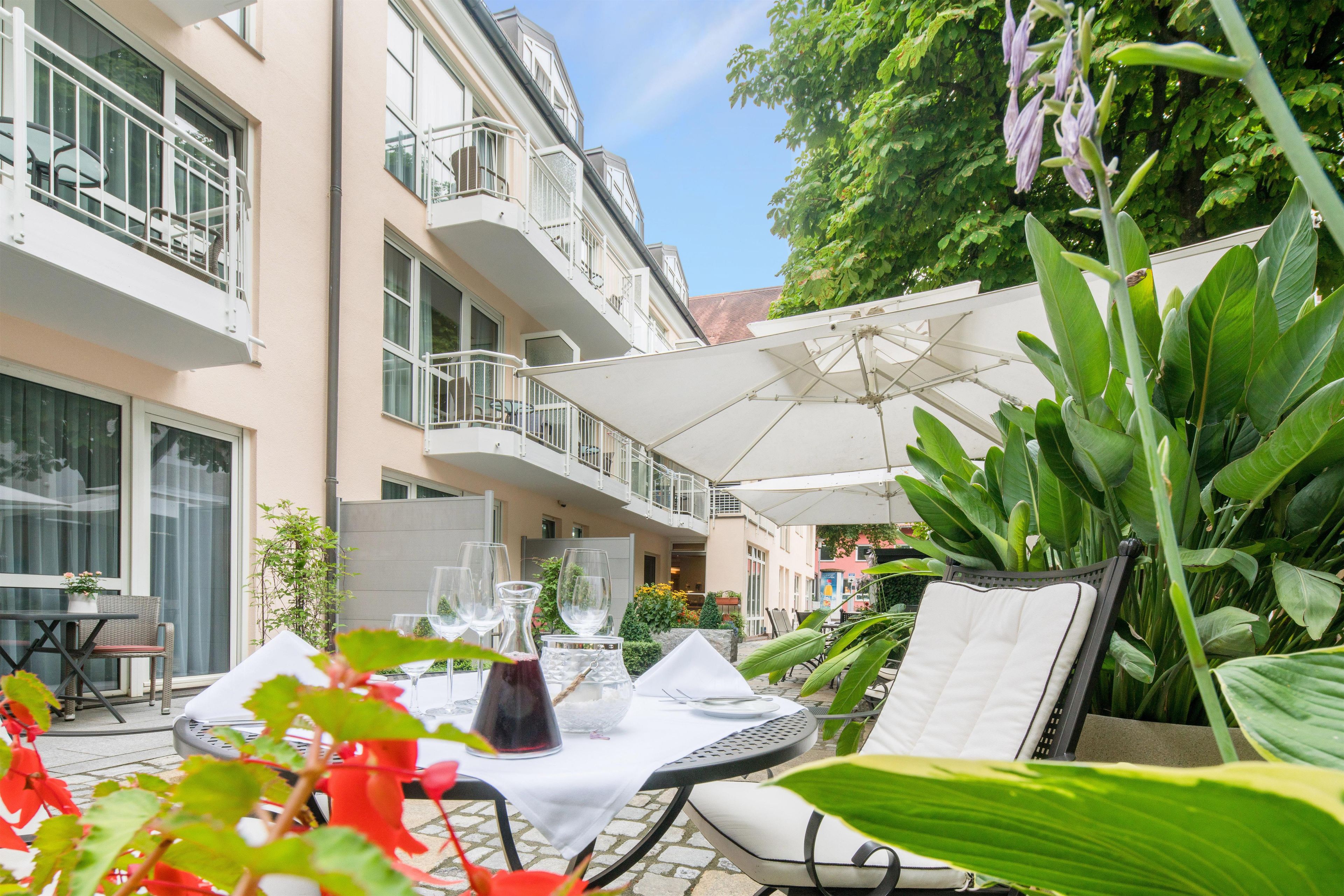 An outdoor seating area with tables and chairs, surrounded by greenery and a building with balconies.