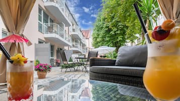 Un área de estar al aire libre con una mesa de vidrio, mobiliario de mimbre y vistas a un edificio de varios pisos con balcones.