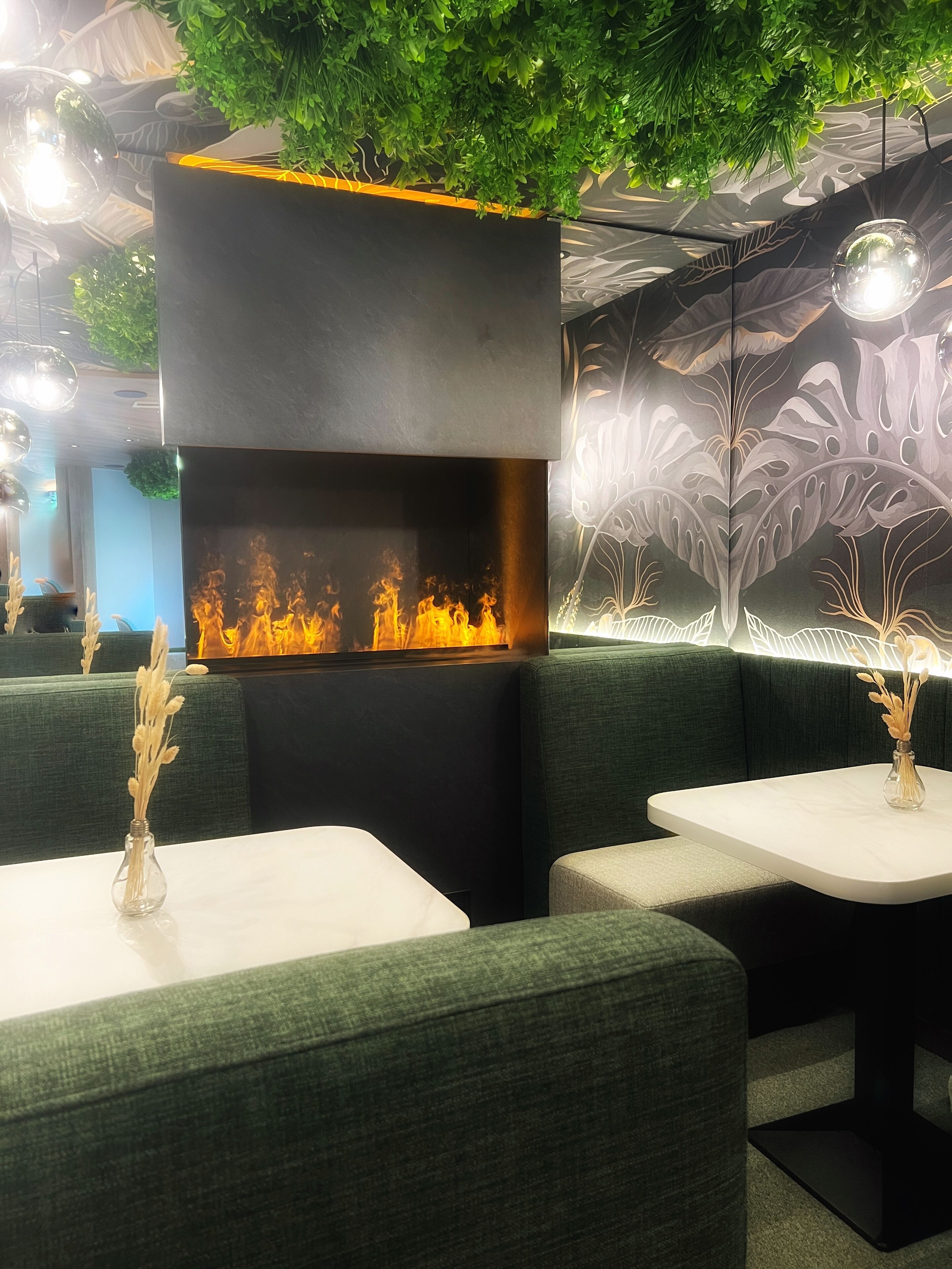 Ein modernes Interieur mit einer zentralen, schwarzen Struktur, die ein Feuerdisplay zeigt, grünen Polstersitzgelegenheiten und dekorativen Vasen auf Tischen.