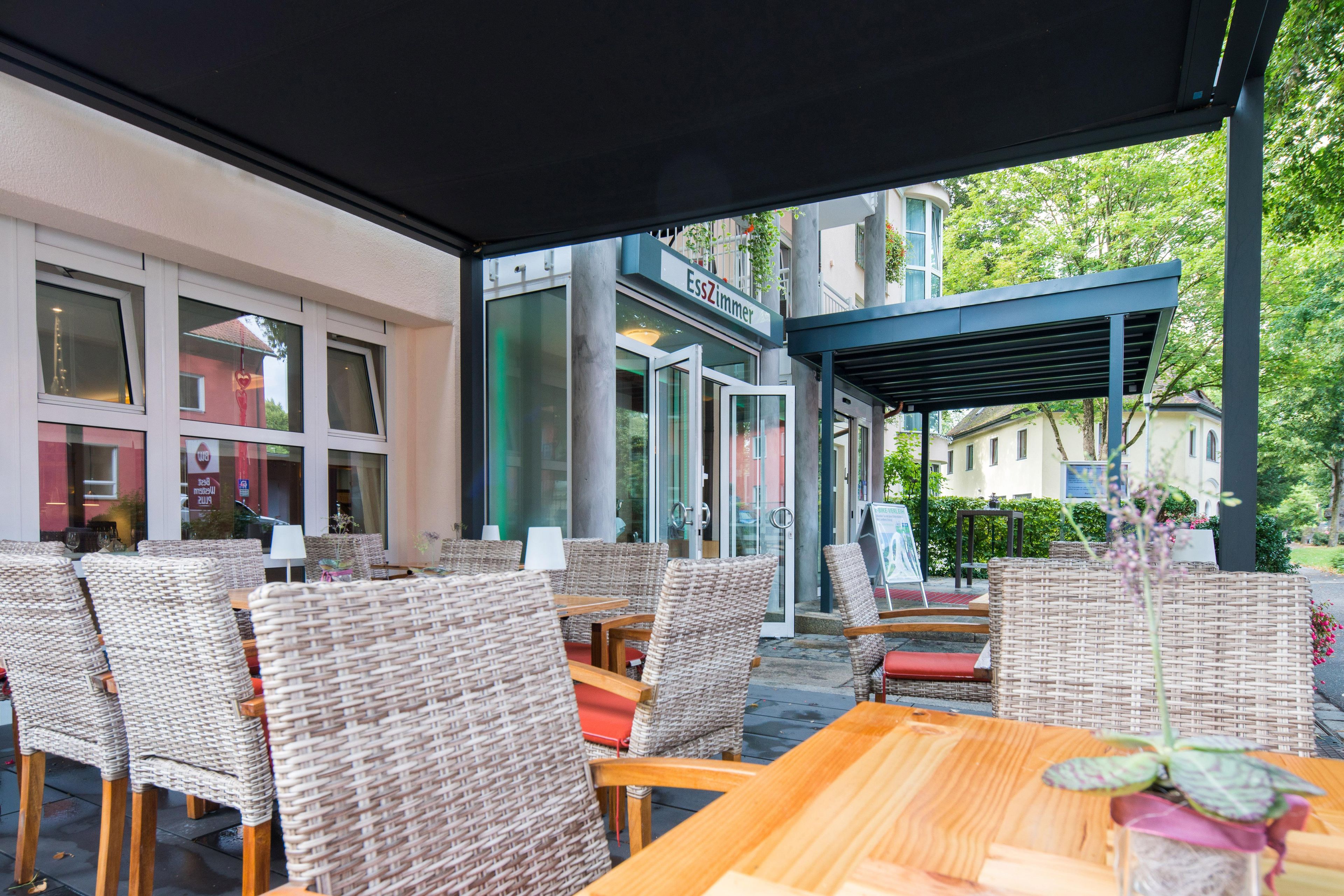 An outdoor seating area with wicker chairs and tables, a covered section, and a building with a sign that reads 'EssZimmer'.