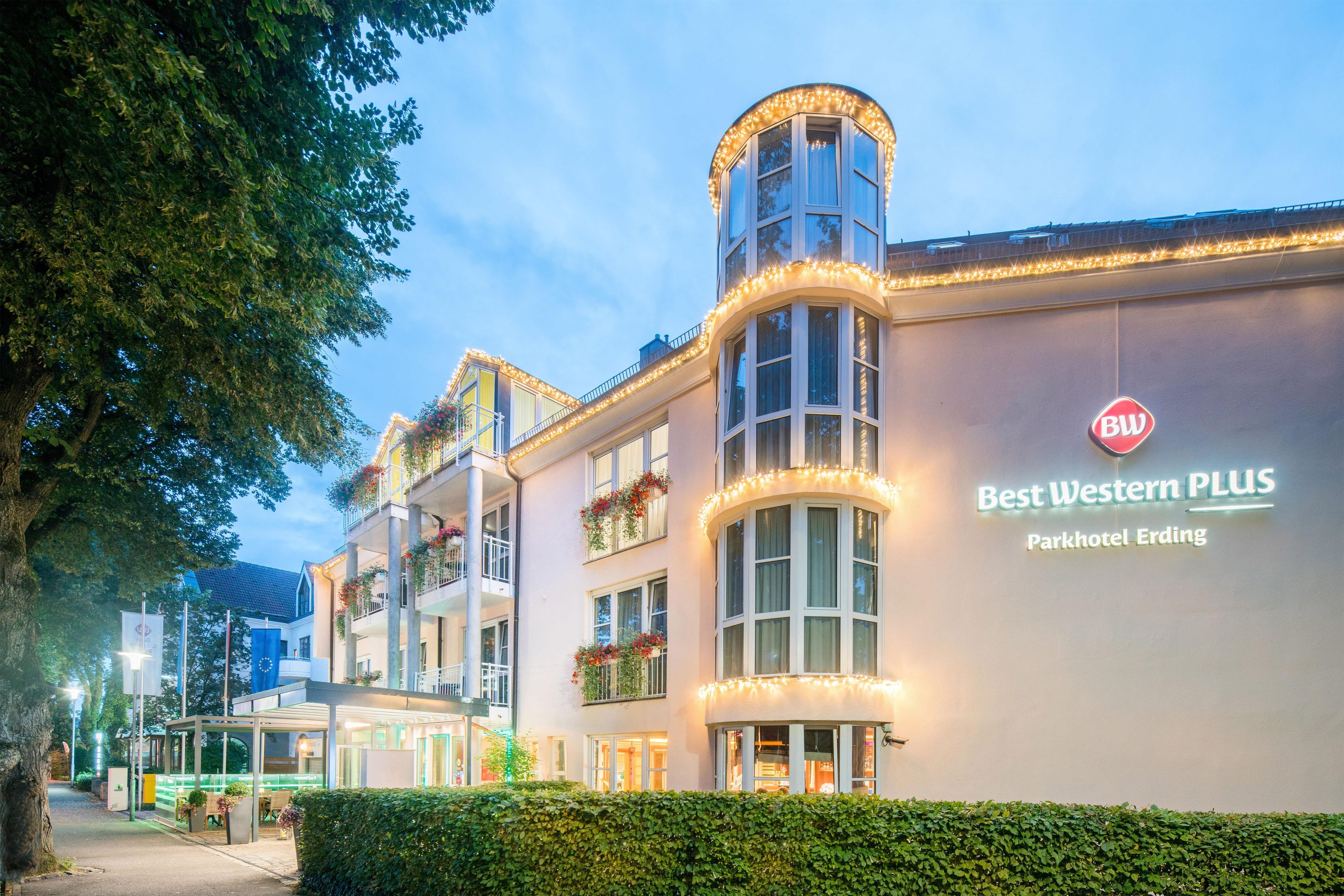 A hotel with a curved corner facade, multiple windows, and a sign indicating it is the Best Western PLUS Parkhotel Erding.
