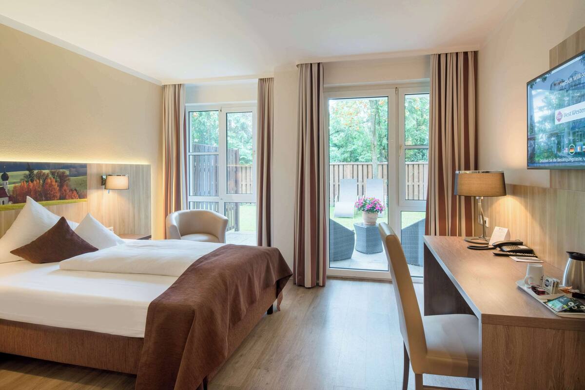 A hotel room with a bed, a desk, a chair, and a balcony with a view of trees and a wooden deck.