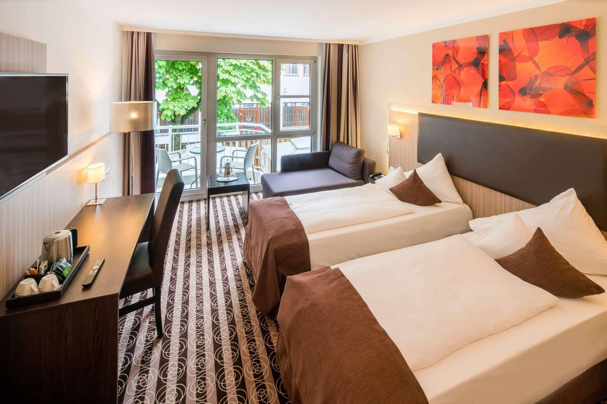 A hotel room with two beds, a desk, a TV, and a balcony with a view of greenery.