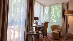 A hotel room with a bed, a desk with a lamp, a chair, a small table with a fruit bowl, and large windows with curtains. - Best Western Plus Parkhotel Erding (Erding)