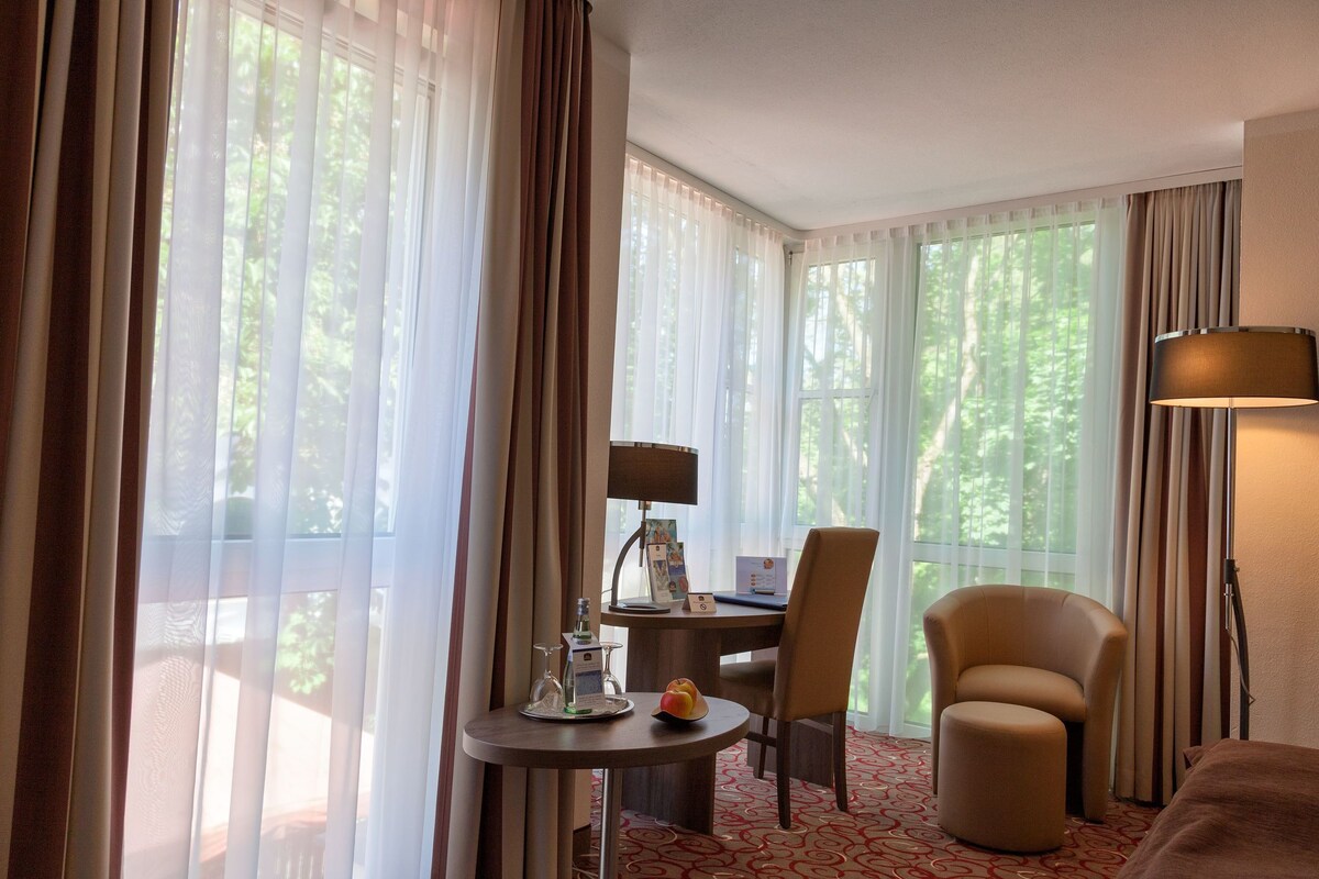 A hotel room with a bed, a desk with a lamp, a chair, a small table with a fruit bowl, and large windows with curtains.