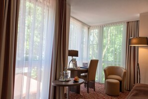 A hotel room with a bed, a desk with a lamp, a chair, a small table with a fruit bowl, and large windows with curtains. - Best Western Plus Parkhotel Erding (Erding)