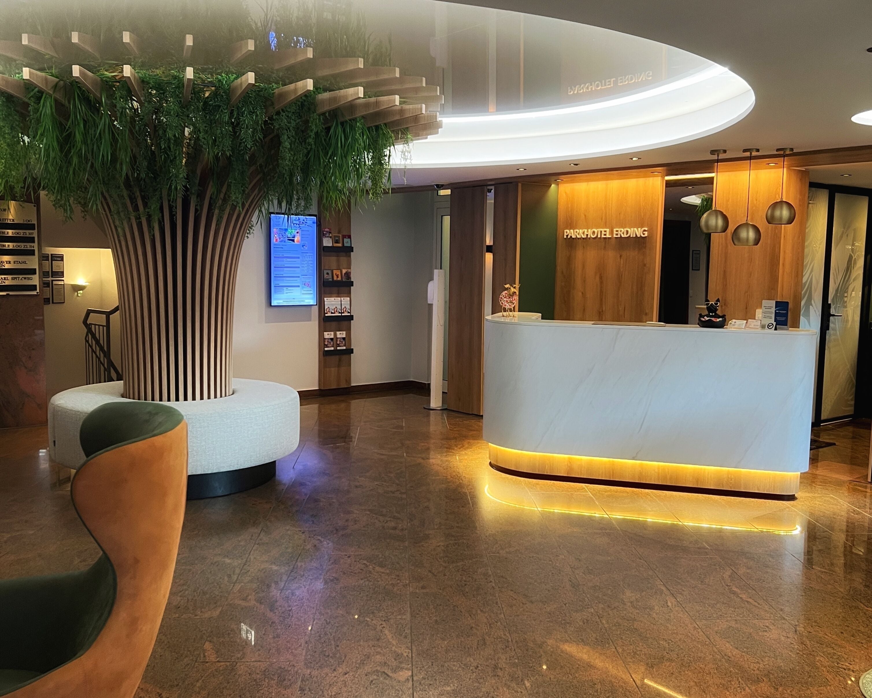 Ein moderner Empfangsbereich mit einem Holzbüro, einer großen Kunstpflanze und einer Sitzecke mit einer gebogenen Bank.