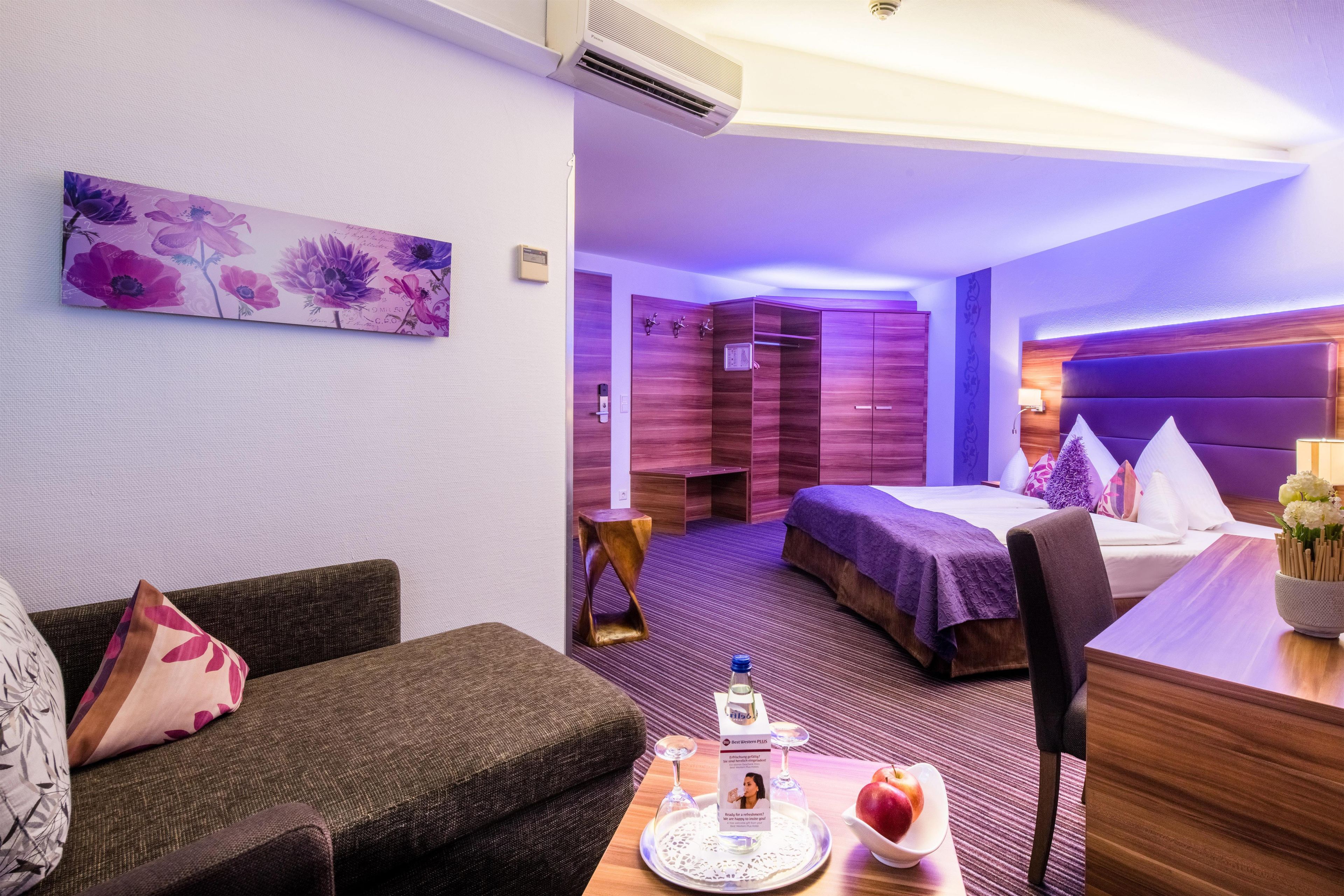 A hotel room with a bed, a sofa, a desk with a tray of apples and a bottle, and a painting on the wall.