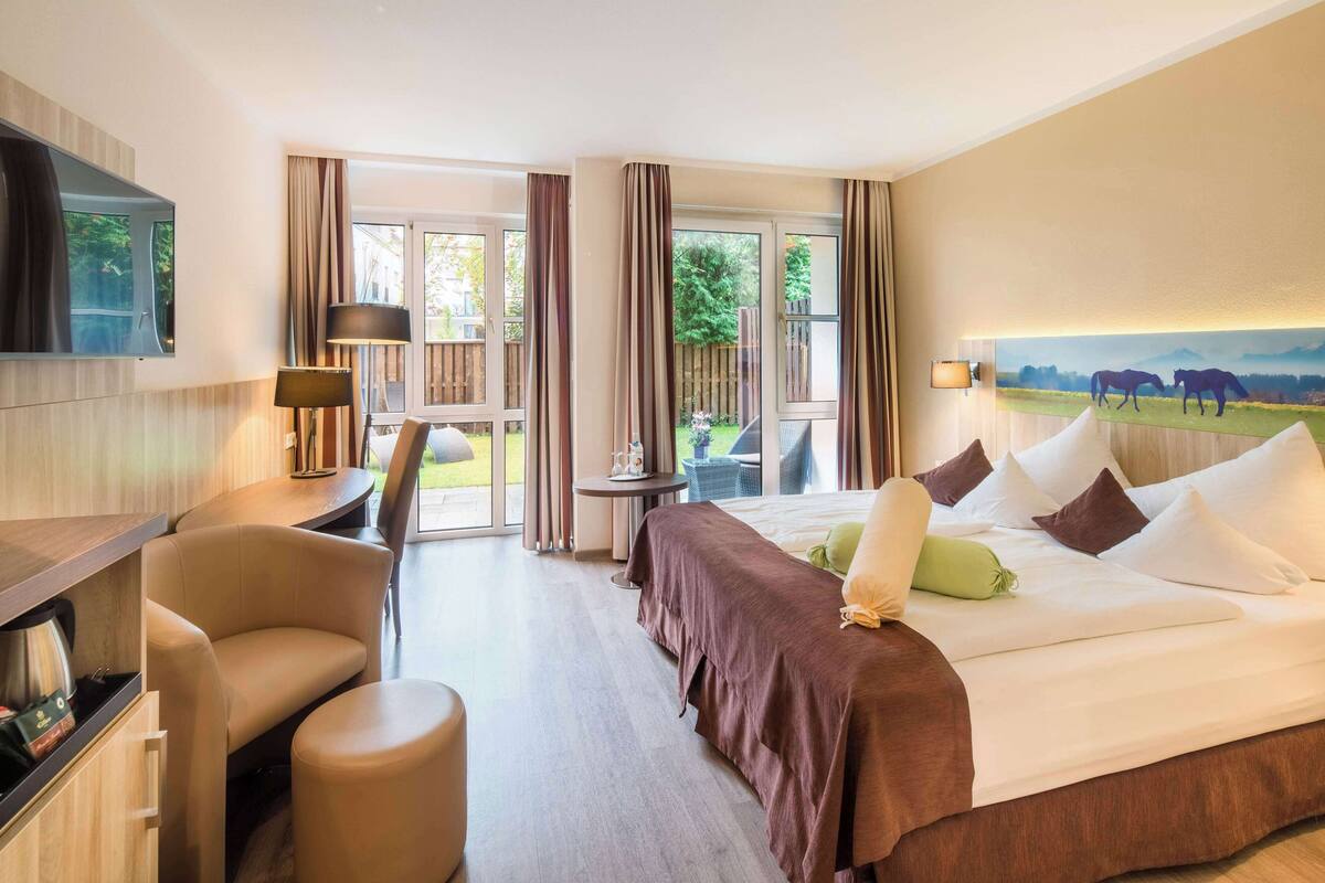 A hotel room with a bed, a desk, a chair, a TV, and a balcony with a view of horses.