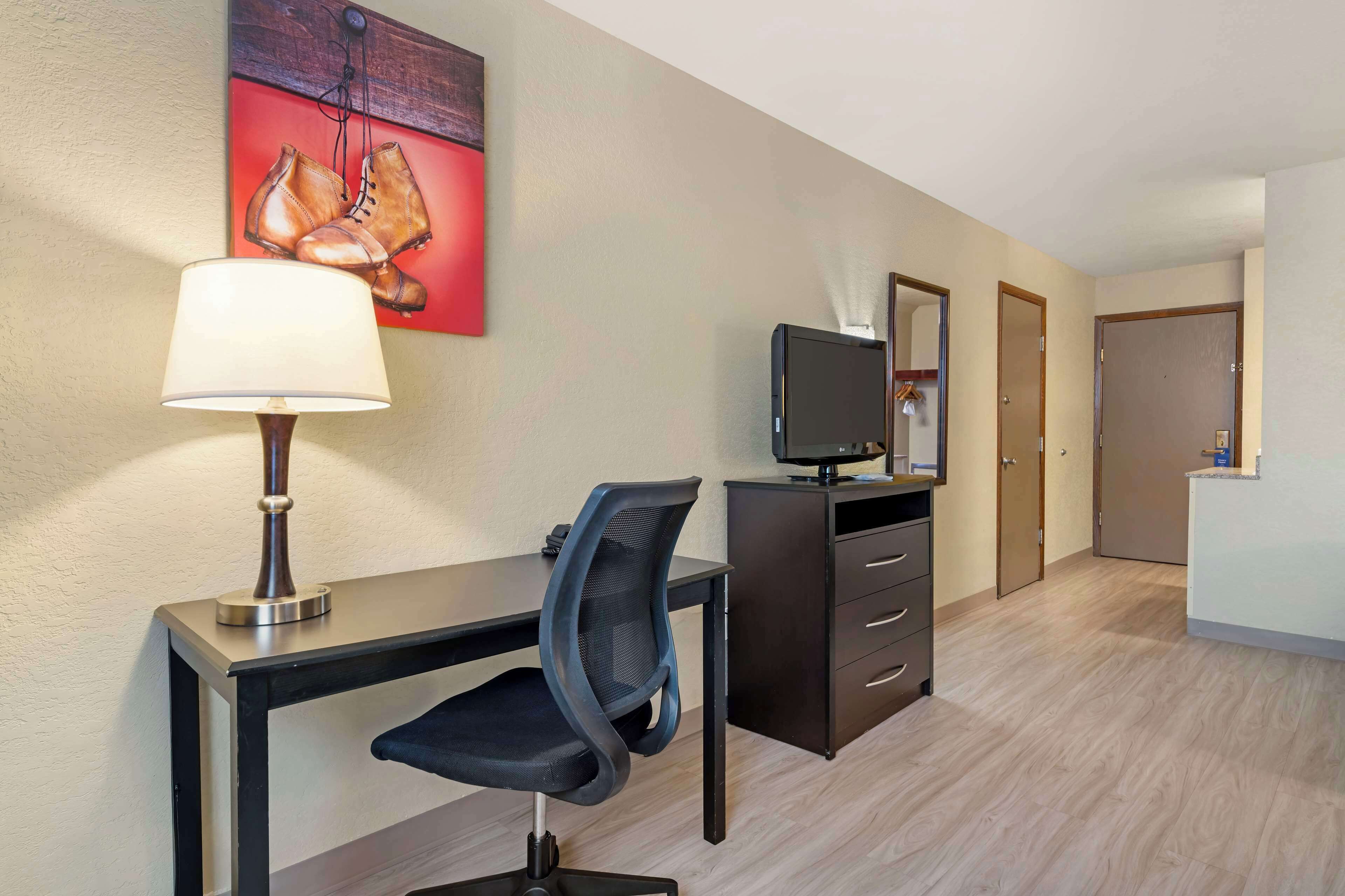 A hotel room with a desk, chair, TV, and a painting of boots on the wall.