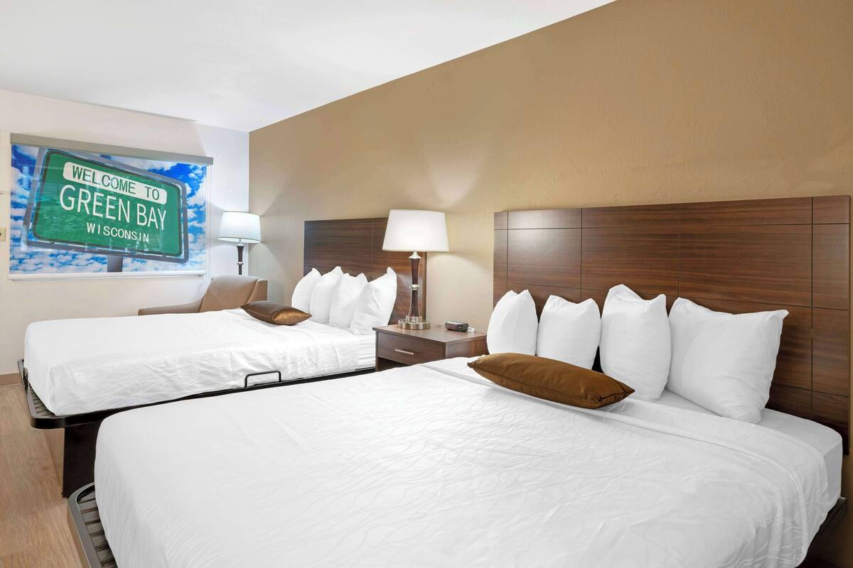 Une chambre d’hôtel avec deux lits, une table de chevet avec une lampe et un panneau d’accueil indiquant « Bienvenue à Green Bay, Wisconsin ».