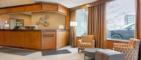 A hotel lobby with a reception desk, patterned armchairs, a small ottoman, and a large window with curtains.